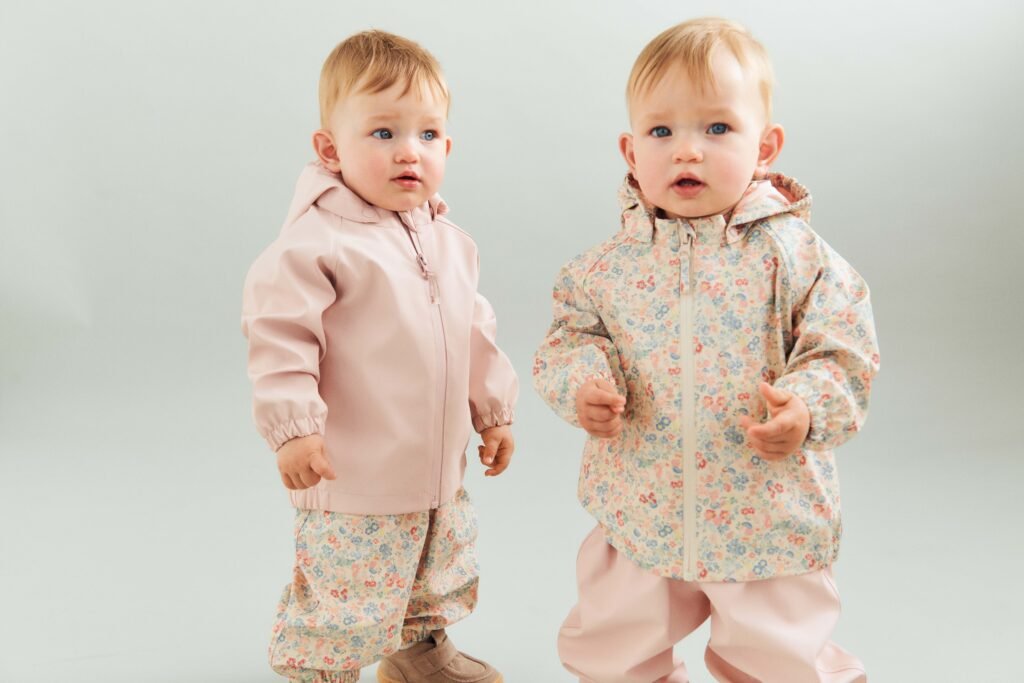Two babies in pastel jackets with floral-pattern pants stand side by side against a pale background.