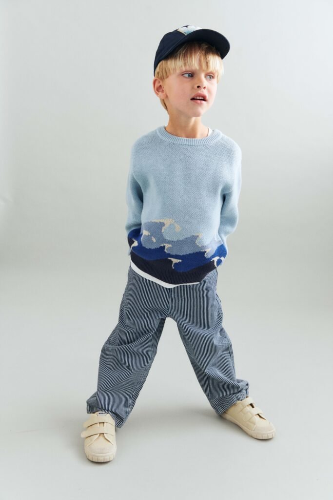 Young boy wearing a light blue knit sweater with wave pattern and striped pants, walking toward the right against a plain background.